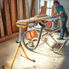 Woodworker feeding long board through table saw with outfeed support stand in workshop