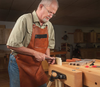 Older man wearing leather apron using chisel to shape wood clamped in woodworking vise