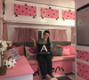 Woman sitting on a pink-painted wooden bench inside a camper with polka dot cabinet doors and decorative cushions.