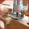 Hands guiding a Bosch plunge router shaping the edge of a wooden board in a workshop.