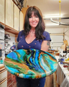 Woman holding a round wooden panel with colorful resin art in a woodworking workshop.