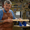 Woodworker wearing leather apron demonstrating holding a wooden board near a Powermatic bandsaw with a blue miter box guide.