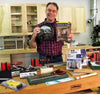 Woodworker in workshop holding a clock and Woodcraft magazine, surrounded by woodworking tools and supplies on a workbench.