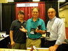 Three men at woodworking booth, one holding a microphone, hand planes visible on table in front