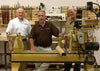 Three men stand around a Powermatic wood lathe with a partially turned wood blank in a woodworking workshop.