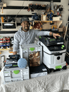 Woodworker smiling next to Festool dust extractor, sander, and toolboxes in workshop setting