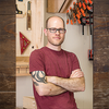 Man with glasses and tattoo standing in workshop with woodworking clamps and tools on wall behind him
