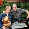Two woodworkers smiling, one holding a carved wooden Santa, the other beside a scroll saw in a workshop setting.