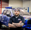 Woodworker with tattooed arms standing in workshop in front of custom painted truck and American flag