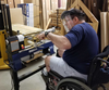 Woodworker in wheelchair using Rikon lathe to shape wood in workshop with stacked lumber.