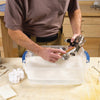 Person cleaning an HVLP spray gun with a brush over a plastic tub filled with soapy water in a woodworking workshop